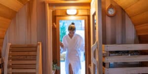 A woman in a white robe stepping out of a wooden sauna with a relaxed expression