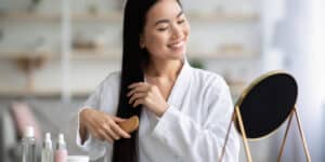 A woman in a white robe smiling while brushing her long hair in front of a mirror