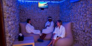Three people in white robes relaxing in a spa room with stone walls and blue lighting